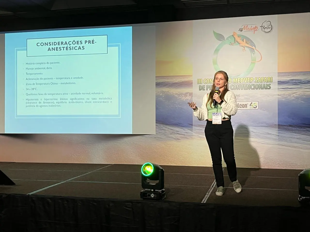 Guta palestrando em um palco, ao lado de um telão com slides sobre considerações pré-anestésicas
